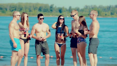 Young-happy-friends-stand-in-a-semicircle-and-communicate-on-the-beach-near-the-water-with-soda-bottles