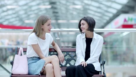 Fade-in-of-two-stylish-girls-resting-on-bench-in-mall-after-shopping,-talking-joyfully-and-laughing