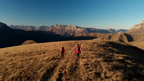 Vista-aérea-de-un-viajero-de-dos-chicas-con-mochilas-y-cámaras-de-pasear-por-las-colinas-entre-las-rocas-épicas-en-las-montañas.-Fotógrafos-de-las-niñas-con-sus-cámaras-al-atardecer