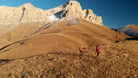 Vista-aérea-de-un-viajero-de-dos-chicas-con-mochilas-y-cámaras-de-pasear-por-las-colinas-entre-las-rocas-épicas-en-las-montañas.-Fotógrafos-de-las-niñas-con-sus-cámaras-al-atardecer