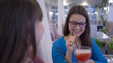 lächelnde-Freundinnen-in-Brillen-Spaß-sagen-und-trinken-Saft-durch-Stroh-während-des-Abendessens-im-Restaurant-im-Urlaub