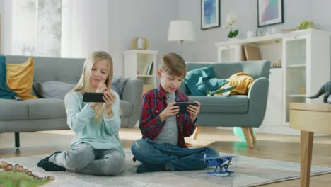 Zu-Hause-auf-einem-Teppich-sitzen:-niedliche-kleine-Mädchen-und-süßen-Boy-spielen-gruppenweise-in-Videospiel-auf-zwei-Smartphones,-hielt-sie-im-horizontalen-Modus-\"Querformat\".-Glückliche-Kinder-tun-High-Five.