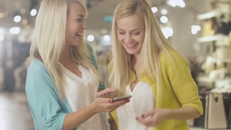 Two-cheerful-blond-girls-are-using-a-smartphone-in-a-department-store.