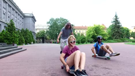 Man-and-woman-sitting-on-a-longboard,-friend-skateboarding-next-to-them