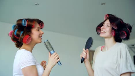 Dos-chicas-divertidas-cantando-con-peines-bailan-y-tienen-alegría-en-la-sala-de-estar-en-casa