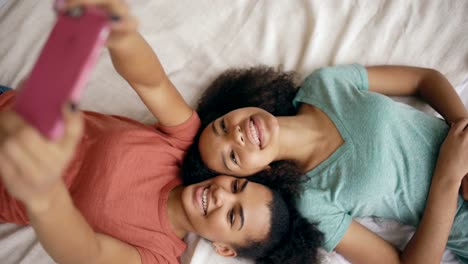 Top-view-of-beautiful-mixed-race-funny-girlfriends-making-selfie-portrait-on-bed-in-bedroom-at-home