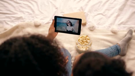 Top-view-of-two-girlfriends-sitting-on-bed-at-home-talking-on-skype-on-tablet-computer-with-their-friend-having-vacation-on-beach