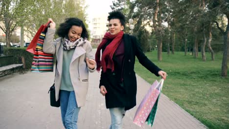 Dolly-Schuss-von-zwei-attraktiven-gemischten-Rennen-Frauen-tanzen-und-Spaß-haben,-während-sie-den-Park-mit-Einkaufstaschen-zu-Fuß.-Glückliche-junge-Freunde-gehen-nach-dem-Besuch-Mall-Verkauf