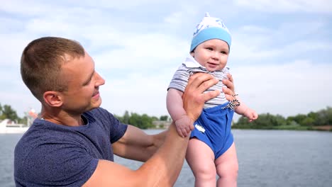 feliz-papá-sostiene-en-primer-hijo-mano-cerca-del-río,-suerte-papá-juega-con-bebé-en-loch,-niño-pequeño-en-las-fuertes-manos-de-padre