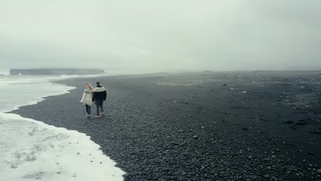 Vista-aérea-de-joven-pareja-caminando-por-la-playa-volcánica-negra-en-Islandia.-Hombre-y-mujer-en-lopapeysa-lanzan-roca-mar