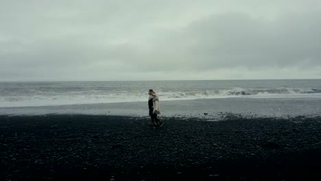 Copter-flying-near-the-young-couple-in-icelandic-sweater.-Man-and-woman-walking,-holding-hands-on-black-volcanic-beach