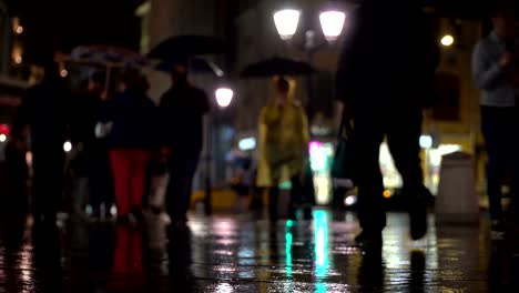 Abstract-background-rainy-late-evening-in-the-city-in-naturale-dark-tones.-Raindrops-fall-on-the-colorful-asphalt-illuminated-by-street-lamps.-Among-the-crowd-of-pedestrians-go-two-friends-under-umbrellas-and-talk.-Lifestyle-of-modern-city