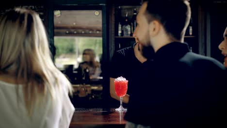 Barman-making-cocktail-for-young-friends-at-the-bar.-A-cheerful-company,-sitting-at-a-bar-with-drinks,-and-talking