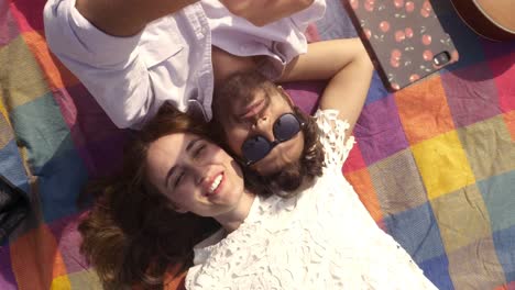 Happy-lovely-young-couple-lying-on-colorful-blanket-in-the-park-taking-selfies-smartphone-romantic-with-guitar-sunglasses-beautiful-attractive-girl-top-view-rotating-camera-slow-motion