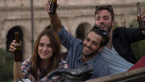 Tres-turistas-jóvenes-amigos-con-bicicletas-sentado-sobre-un-banco-frente-a-Coliseo-bajo-el-árbol-en-la-puesta-de-sol-beber-cervezas-diversión-hablando-riendo-a-refrigeración-en-cámara-lenta-de-Roma
