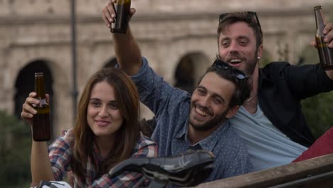 Three-young-friends-tourists-with-bikes-sitting-on-bench-in-front-of-colosseum-under-tree-at-sunset-drinking-beers-having-fun-talking-laughing-chilling-in-Rome-slow-motion