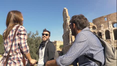 Three-young-friends-tourists-sitting-in-front-of-colosseum-in-rome-talking-having-fun-near-arch-of-constantine-with-backpacks-sunglasses-happy-beautiful-girl-long-hair