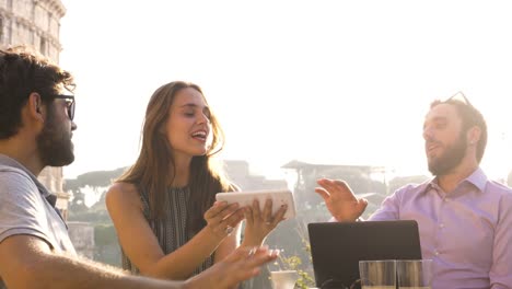 Drei-junge-Menschen,-die-gemeinsam-an-einem-Projekt-mit-Laptop-und-Tablet-Brainstorming-schreiben,-sprechen-und-die-Erforschung-neuer-Ideen,-die-sitzen-am-Stehtisch-Restaurant-vor-Colosseum-in-Rom-bei-Sonnenuntergang