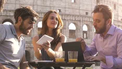 Drei-junge-Menschen,-die-gemeinsam-an-einem-Projekt-mit-Laptop-und-Tablet-Brainstorming-schreiben,-sprechen-und-die-Erforschung-neuer-Ideen,-die-sitzen-am-Stehtisch-Restaurant-vor-Colosseum-in-Rom-bei-Sonnenuntergang