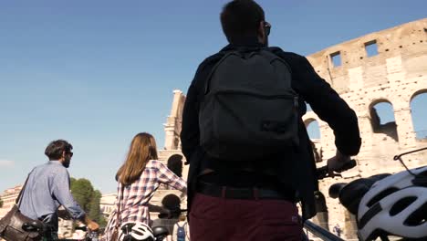 Drei-junge-Freunde-Touristen-zu-Fuß,-mit-Fahrrad-und-Rucksack-im-Colosseum-in-Rom-am-sonnigen-Tag-Zeitlupe-Kamera-Steadycam