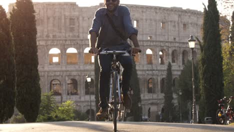 Tres-turistas-de-jóvenes-amigos,-montar-en-bici-en-el-Parque-colle-oppio-frente-Coliseo-en-carretera-con-árboles-al-atardecer-en-Roma-lenta