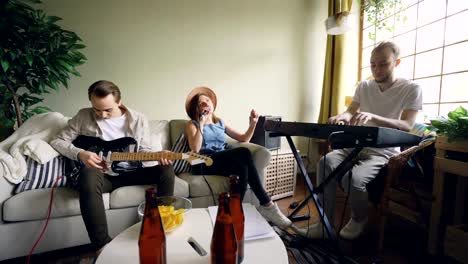 Two-cheerful-guys-musicians-are-playing-keyboard-and-guitar-while-their-female-friend-vocalist-is-singing-holding-microphone.-Friendship-and-music-concept.