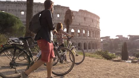 Drei-glückliche-junge-Freunde-Touristen-mit-Fahrrädern-und-Rucksäcke-am-Kolosseum-in-Rom-ankommen-auf-Hügel-bei-Sonnenuntergang-mit-Bäumen-Zeitlupe-steadycam