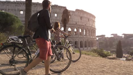 Three-happy-young-friends-tourists-with-bikes-and-backpacks-at-Colosseum-in-Rome-arriving-on-hill-at-sunset-with-trees-slow-motion-steadycam