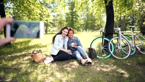 Friend-is-photographing-two-pretty-girls-sitting-on-lawn-in-park-using-phone-holding-it-in-hand,-focus-on-smartphone-screen-then-on-people.-Portrait-and-technology-concept.