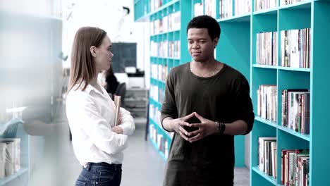 Tiro-de-seguimiento-de-muchacha-caucásica-y-africano-niño-hablando-en-la-biblioteca-de-la-Universidad-cerca-de-estantes