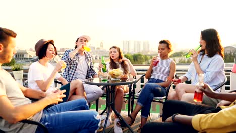 Jóvenes-alegres-celebran-fiesta-del-techo-beber-y-tintineantes-botellas-y-vidrios-entonces-hablando-y-riendo.-Hermosa-vista-de-la-ciudad-moderna-está-en-el-fondo.