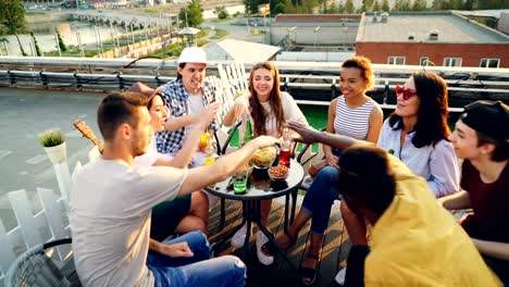 Happy-friends-are-putting-hands-together-then-raising-them-celebrating-success-sitting-at-table-on-rooftop,-laughing-and-talking.-Friendship-and-party-concept.