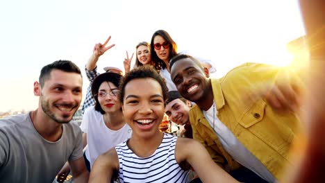 Punto-de-vista-toma-de-muchacha-afroamericana-con-cámara-y-tomando-selfie-con-amigos-felices-en-la-fiesta-en-la-azotea.-Hombres-y-mujeres-están-mirando-a-cámara,-posando-y-riendo.