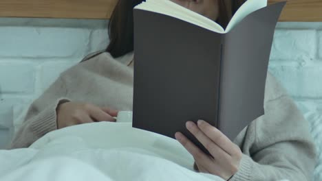 Hermosa-mujer-asiática-disfruta-de-beber-café-caliente-y-lectura-en-cama-en-su-dormitorio.-Mujer-de-Asia-usando-suéter-cómodo-sosteniendo-un-libro-y-una-taza-de-coffee.lifestyle-mujer-de-asia-en-el-concepto-de-hogar