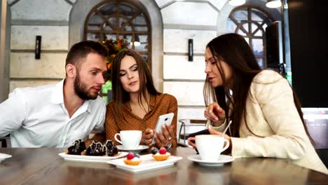 Eine-Gruppe-von-Männern-und-Frauen-im-Büro-Kleidung-sitzen-in-einem-Café-an-einem-großen-Tisch-und-zeigen-einander-in-Mobile-Handys-lustige-Bilder