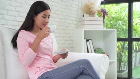 Mujer-asiática-joven-alegre-beber-café-caliente-o-té-disfrutando-mientras-está-sentado-en-su-sala-de-estar-en-casa.-Atractiva-mujer-asiática-feliz-sosteniendo-una-taza-de-café.