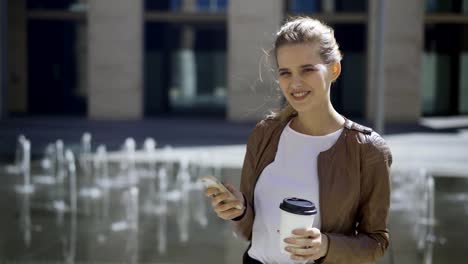 Schöne-blonde-Frau-mit-Einweg-Kaffeetasse-und-Handy-in-den-Händen-posiert-in-der-Nähe-von-Brunnen-in-der-Straße-und-glücklich-lächelnd-in-die-Kamera,-mittlere-Schuss