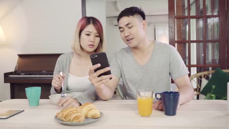 Attractive-young-Asian-couple-distracted-at-table-with-newspaper-and-cell-phone-while-eating-breakfast.-Excited-young-Asian-couple-surprised-by-unbelievably-good-news,-happy-family-amazed-by-internet.