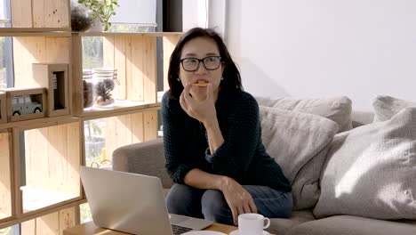 Hermosa-joven-de-los-asiáticos-trabajando-con-ordenador-portátil-y-comer-meriendas,-tomando-café-sentado-en-el-sofá-en-casa.-concepto-de-trabajo-en-casa