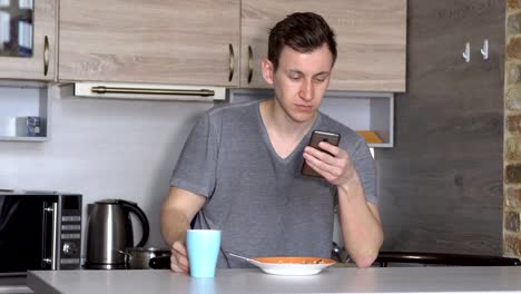 Junger-Mann-mit-einem-Telefon-frisst-an-einem-Tisch-in-der-Küche