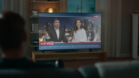 Man-Watches-Late-Night-News-Show-on-TV-while-Sitting-on-a-Couch-at-Home-in-the-Evening.-Two-Presenters-Talk-and-Joke-on-TV.-Cozy-Living-Room-with-Warm-Lights.-Over-the-Shoulder-Shot.