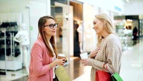 Cute-girls-friends-are-chatting-and-laughing-in-shopping-center-holding-takeout-drinks-and-colorful-paper-bags.-Joyful-conversation,-good-mood-and-youth-concept.