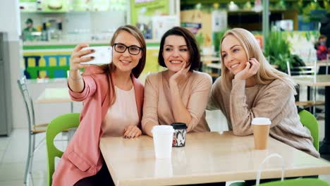 Schöne-junge-Frauen-nehmen-Selfie-mit-Smartphone-am-Tisch-im-Café-sitzen-und-posiert-mit-Getränken.-Freundschaft,-Freizeit-und-moderne-Technologie-Konzept.