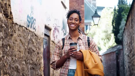 Joven-mujer-negra-con-un-pie-de-camisa-a-cuadros-en-un-callejón-con-su-capa-y-un-café-para-llevar,-sonriendo-a-la-cámara,-de-cerca