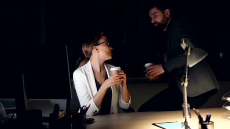 Cansado-oficinista-joven-trabaja-con-ordenador-por-la-noche-cuando-su-compañero-de-trabajo-lleva-su-café-para-llevar.-Personas-tienen-conversación-amistosa.