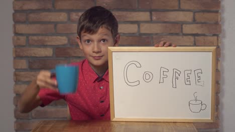 Kind-am-Schreibtisch-hält-Flipchart-mit-Schriftzug-Kaffee-auf-dem-Hintergrund-roten-Backsteinmauer