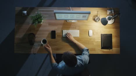 Top-View-of-Young-Creative-Designer-Working-at-His-Desktop-Computer-at-Night.-He-Drinks-Coffee.-Table-Light-Turned-On.