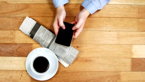 Businesswoman-using-mobile-phone-at-desk-with-newspaper-and-cup-of-coffee-on-table