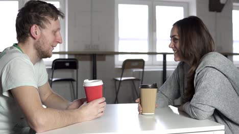 Two-friends-are-sitting-at-the-table-in-cafe,-drinking-coffee-to-go-in-paper-cups-and-talking.-Shot-in-4k