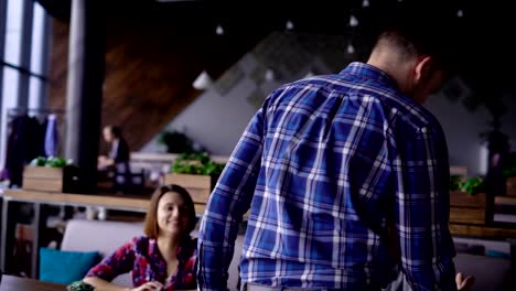 A-middle-aged-man-walks-to-the-restaurant-table,-where-his-friends-sit-and-says-a-welcome,-then-sits-on-a-chair,-his-girlfriend-waving-to-him,-she-is-happy-to-see-a-man